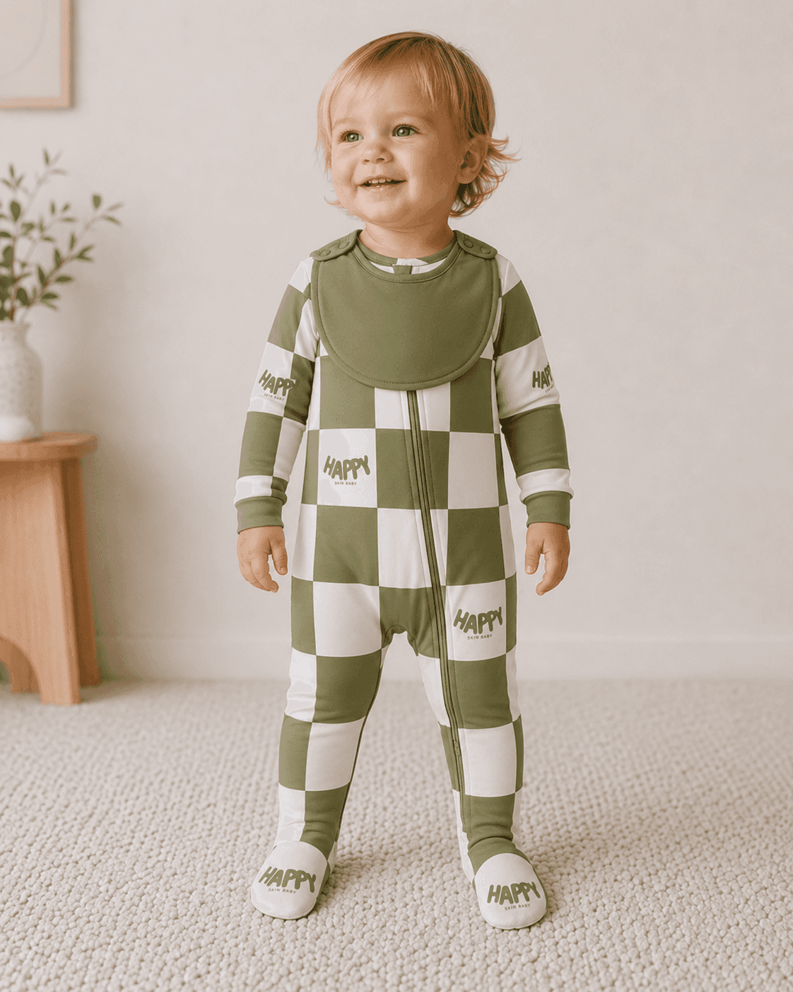 Smiling toddler standing in Happy Skin Baby's olive checker footed pajama with the new attached snap-on bib